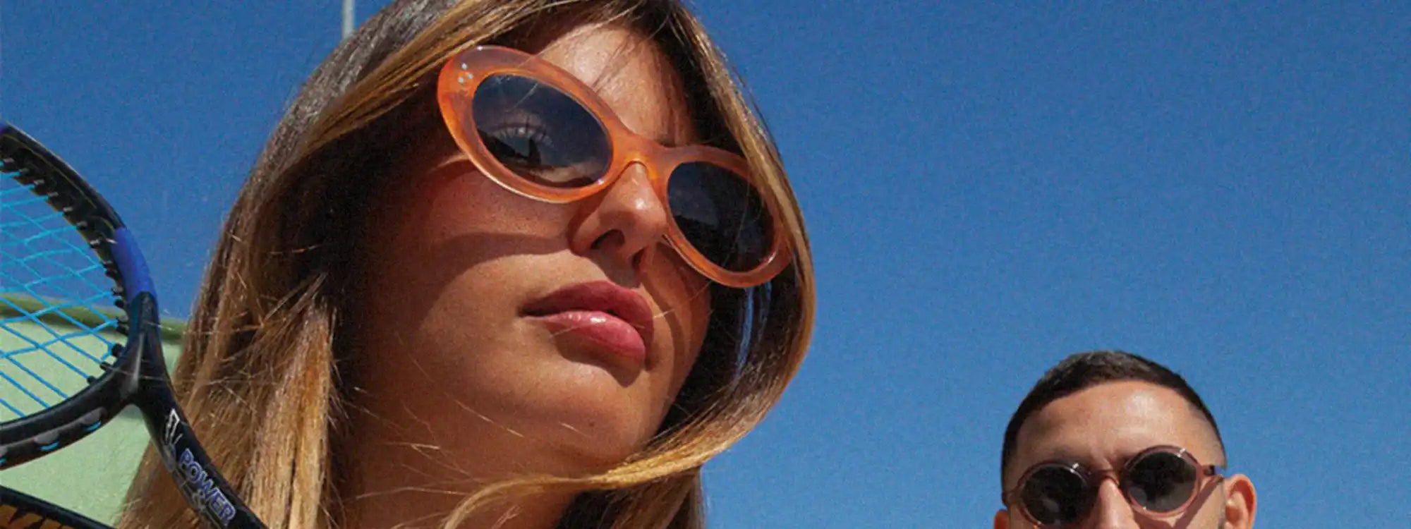 Une femme portant des lunettes de soleil oranges ovales et tenant une raquette de tennis, avec un homme partiellement visible en arrière-plan.