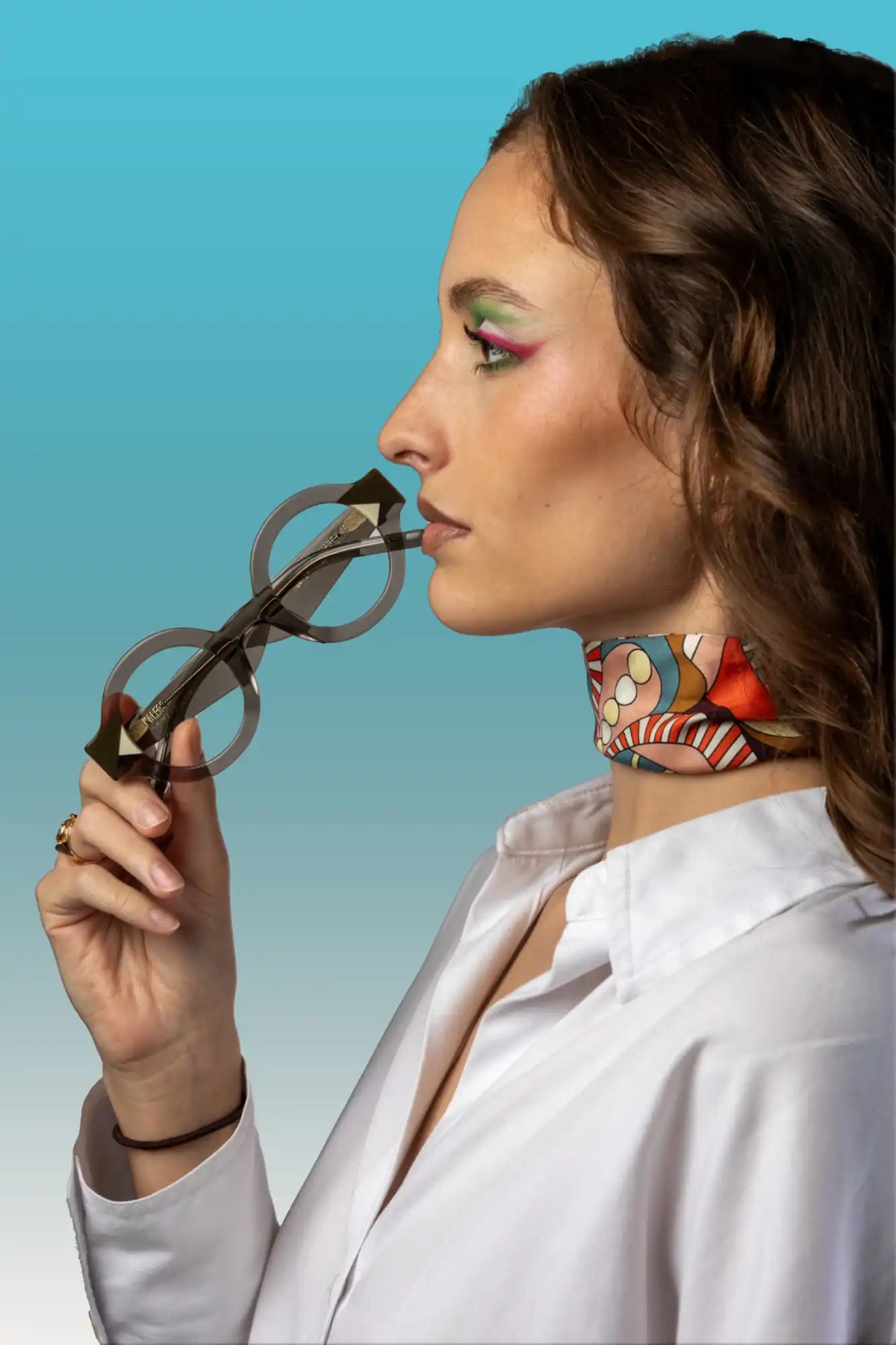 Une femme aux longs cheveux bruns, portant une chemise blanche et un foulard coloré au cou, tenant une paire de lunettes rondes dans sa main et près de sa bouche.