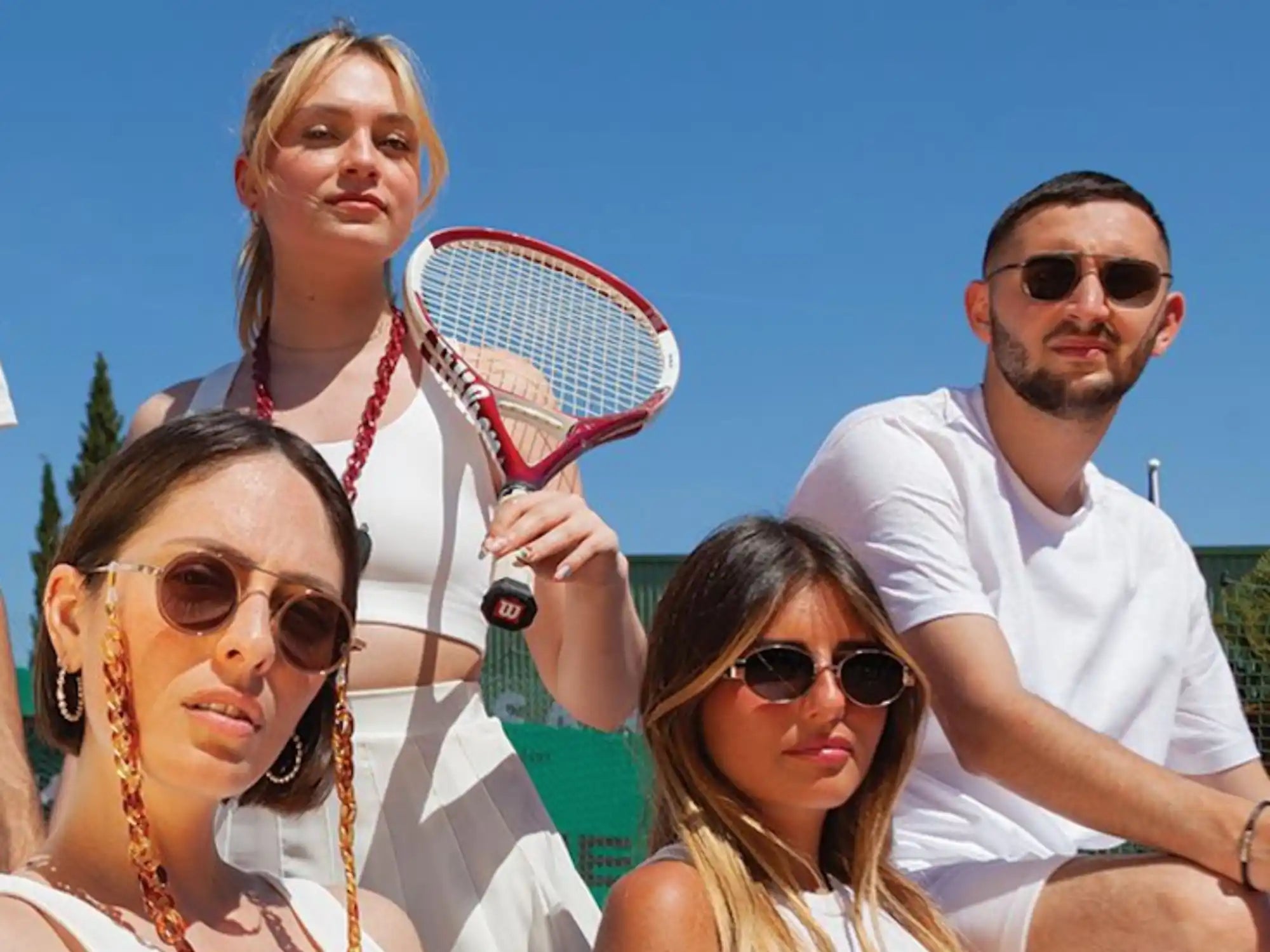 Un groupe de quatre jeunes adultes portant des lunettes de soleil et des tenues blanches, l'un tenant une raquette de tennis rouge et blanche.